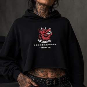 May include: Black cropped hoodie with a red and white graphic of a snarling dog's head. The text "UNDERGROUND TRADING CO." is printed below the graphic in white. The hoodie has a drawstring hood and long sleeves.