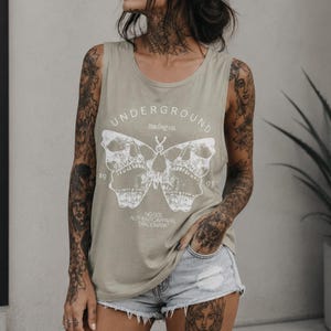 Puede incluir: Camiseta sin mangas verde oliva con un diseño de mariposa blanca, con detalles de calavera. El texto "UNDERGROUND trading co." y "19 93" están impresos encima y debajo de la mariposa. La parte inferior de la camiseta dice "NO.001 AUTHENTIC APPAREL TRADEMARK."