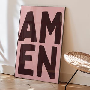 Könnte beinhalten: Ein gerahmter Kunstdruck mit dem Wort "AMEN" in dunkelbraunen Blockbuchstaben auf hellrosa Hintergrund. Das Kunstwerk lehnt an einer weißen Wand auf einem Holzboden, mit einem Teil eines Korbstuhls sichtbar.
