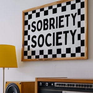 Puede incluir: Una obra de arte enmarcada con las palabras "SOBRIETY SOCIETY" en letras negras sobre un fondo a cuadros blanco y negro. Una lámpara amarilla y un equipo estéreo de madera se encuentran debajo de la obra en un estante blanco.