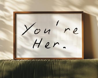 May include: A framed print with the handwritten text "You're Her" in black against a white background. The frame is brown, and the print rests above a green velvet sofa. Sunlight casts shadows on the wall.