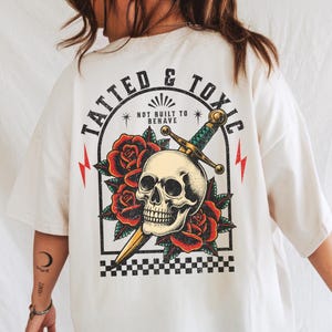 May include: Cream-colored t-shirt with a graphic featuring a skull, roses, and a sword. The text "TATTED & TOXIC" arches above the design, with "NOT BUILT TO BEHAVE" below. Red lightning bolts frame the design.