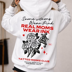 May include: White hoodie with the text "Some Moms Wear Pink REAL MOMS WEAR INK" in black and red. The back of the hoodie features a tattoo gun, roses, and the words "TATTED MOMS CLUB" and an address.