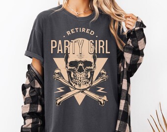 Puede incluir: Una camiseta gris oscuro con un gráfico de calavera y huesos cruzados. El texto "Retired Party Girl" está impreso en la camiseta.