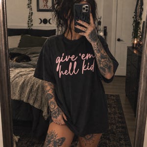 Puede incluir: Una camiseta de manga corta gris carbón con el texto "give 'em hell kid" impreso en una fuente cursiva de color rosa claro en el pecho. La prenda tiene cuello redondo y un corte holgado, confeccionada en tela de algodón suave y lavada.