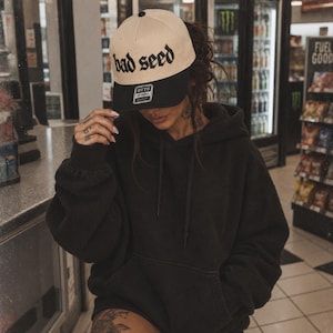 Pode incluir: Uma camisola preta com capuz, mangas compridas e bolso canguru, combinada com um boné de basebol bicolor. O boné apresenta uma coroa cor de creme com o texto "bad seed" em letras góticas pretas e uma aba preta sólida.