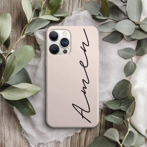 May include: A light pink phone case with the word "Amen" in black script. The case has a camera cutout and is surrounded by green leaves and a white cloth on a wooden surface.