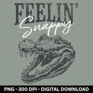 May include: A digital graphic design on a sage green background featuring a black distressed illustration of an alligator head with its mouth wide open. Above the alligator, the text reads "FEELIN'" in a bold, dark, textured font and "Snappy" in a white, cursive script font.