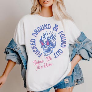 Puede incluir: Sudadera blanca con un gráfico azul y rosa de una calavera con llamas saliendo de sus ojos. El texto alrededor de la calavera dice "F*cked Around & Found Out". Debajo de la calavera está el texto "Sober 'Till It's Over".