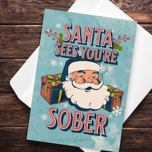May include: A Christmas greeting card with a vintage design. The card features Santa Claus with the text "SANTA SEES YOU'RE SOBER" in red and white lettering. The background is light blue with snowflakes and gift boxes.