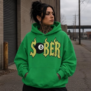 May include: A bright green hooded sweatshirt featuring a graphic on the chest with the word SOBER in a yellow flame-style font, where the letter O is replaced by a black eight ball with a white number eight in the center, and a front pocket.