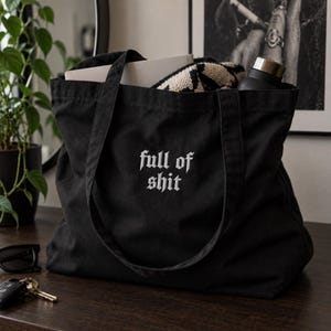 May include: A black canvas tote bag featuring the white embroidered text full of shit in a gothic font. The bag has two shoulder straps and is filled with a white rectangular object, a black and white patterned textile, and a black water bottle.