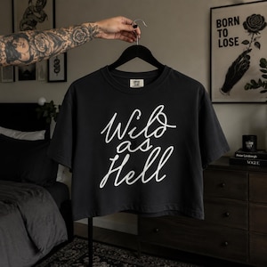 May include: Black crop top with the white text "Wild as Hell" in a handwritten style. The shirt is hanging on a black hanger. The shirt is a crew neck style.