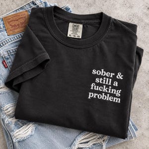Op de afbeelding: Een zwart t-shirt met korte mouwen en een ronde hals, voorzien van de witte tekst "sober & still a fucking problem" aan de rechteronderkant. Het shirt heeft een Comfort Colors merklabel in de nek met de maatvermelding XL-TG-EG.