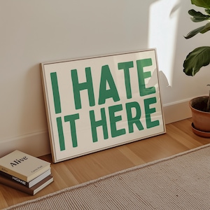 May include: A white framed print with the text "I HATE IT HERE" in green letters on a cream background.