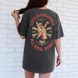 Puede incluir: Camiseta gris oscuro con un gráfico desgastado en la espalda. El gráfico presenta una mano sosteniendo una daga, huesos cruzados y el texto "I Hold Grudges, All Heal in Hell". El texto adicional incluye "Palermo City Sicily", "Raging Misfits", "Hell Bound", "Brooklyn New York".