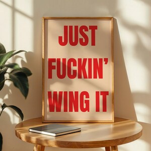 以下が含まれることがあります： ベージュの背景に赤い文字で「JUST FUCKIN' WING IT」と書かれたフレーム入りのアートプリント。明るい茶色の木製フレームに入っており、丸い木製のテーブルの上に置かれています。テーブルの上には閉じられたノートがあります。