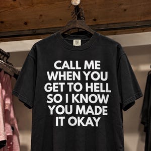 Könnte beinhalten: Ein schwarzes Kurzarm-T-Shirt mit dem weißen Aufdruck CALL ME WHEN YOU GET TO HELL SO I KNOW YOU MADE IT OKAY. Das Kleidungsstück hat einen Rundhalsausschnitt und hängt an einem dunklen Kleiderbügel, was ein schlichtes Grafikdesign zeigt.