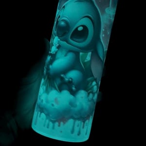 May include: A cylindrical tumbler featuring a teal illustration of the character Stitch from Lilo & Stitch. The design includes Stitch on a cloud, with a space-themed background. The tumbler glows in the dark, creating a vibrant effect.
