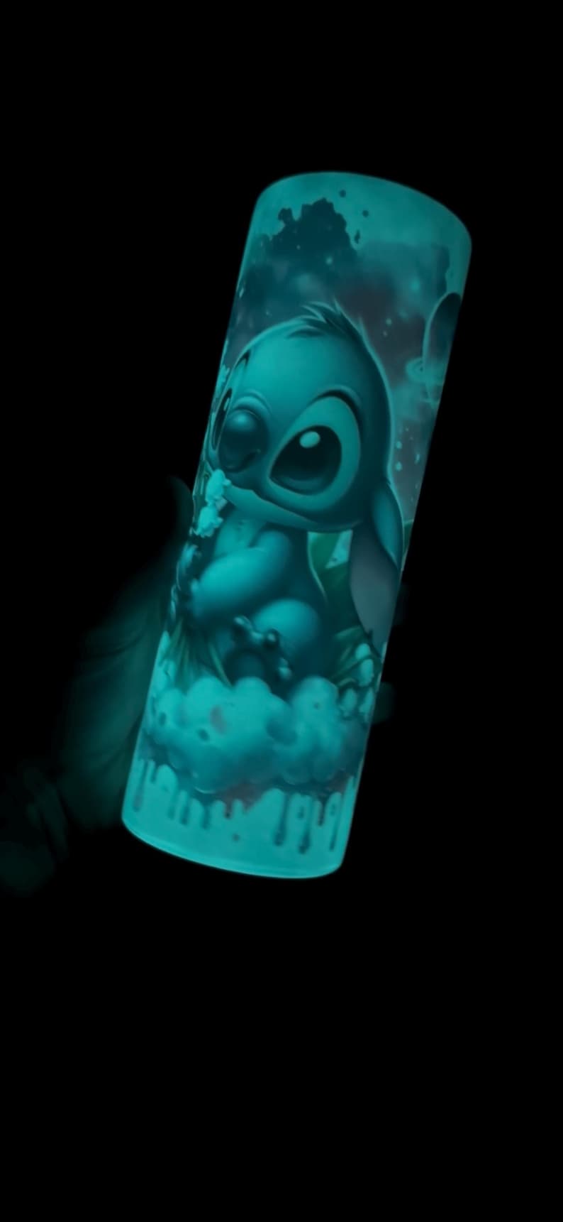 May include: A cylindrical tumbler featuring a teal illustration of the character Stitch from Lilo & Stitch. The design includes Stitch on a cloud, with a space-themed background. The tumbler glows in the dark, creating a vibrant effect.