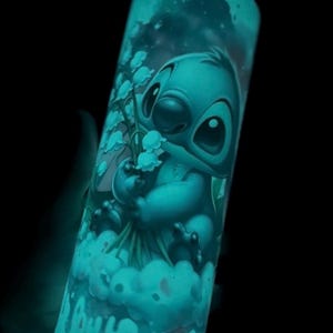 May include: A teal-colored tumbler featuring a cartoon illustration of Stitch holding flowers. The design glows in the dark, with a cloud and starry sky background. The tumbler is cylindrical and appears to be made of glass or a similar material.