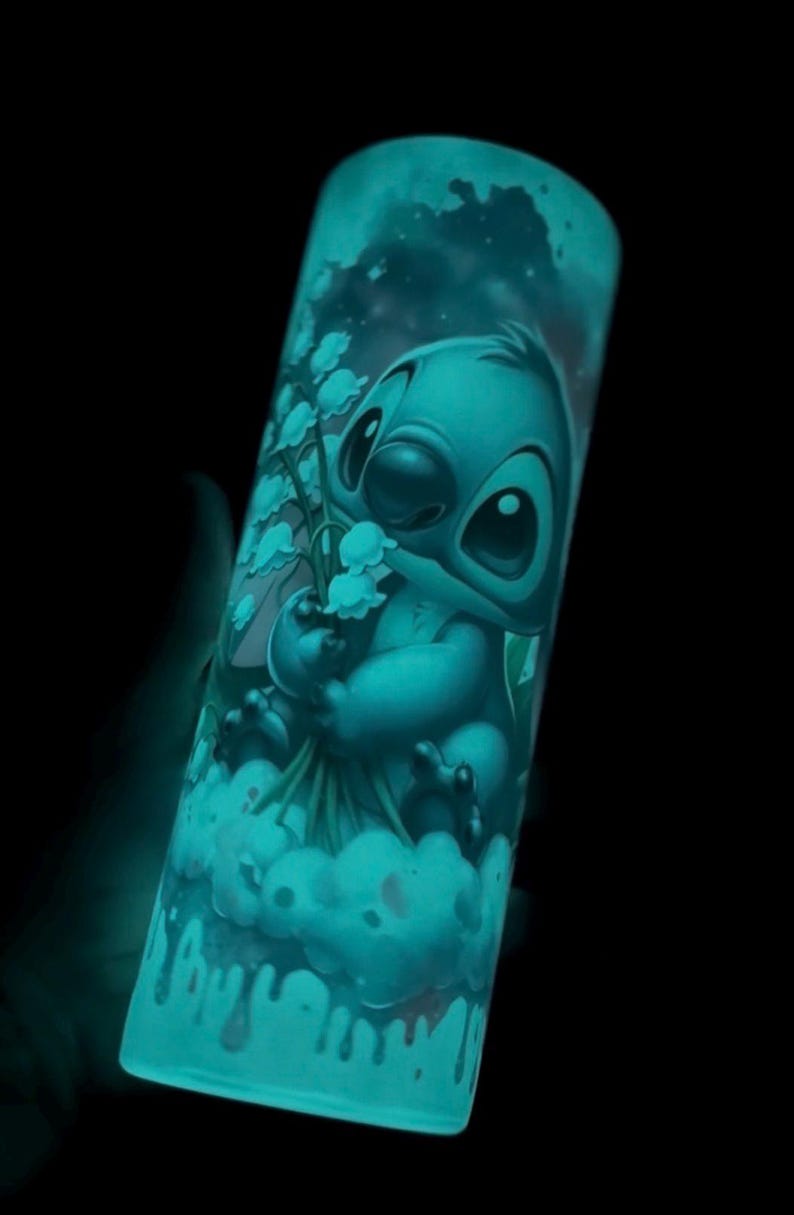May include: A teal-colored tumbler featuring a cartoon illustration of Stitch holding flowers. The design glows in the dark, with a cloud and starry sky background. The tumbler is cylindrical and appears to be made of glass or a similar material.