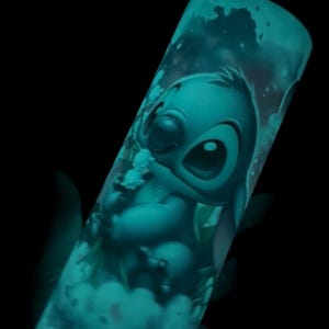 May include: A teal-colored tumbler featuring a cartoon illustration of Stitch from Lilo & Stitch. The design glows in the dark, showcasing Stitch's face and form against a cloudy background. The tumbler is cylindrical and appears to be made of plastic.