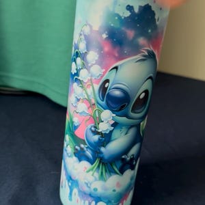 May include: A blue tumbler featuring the character Stitch from Lilo & Stitch, holding white flowers. The design includes a sky-blue background with pink and blue accents. The tumbler is cylindrical and appears to be made of metal.