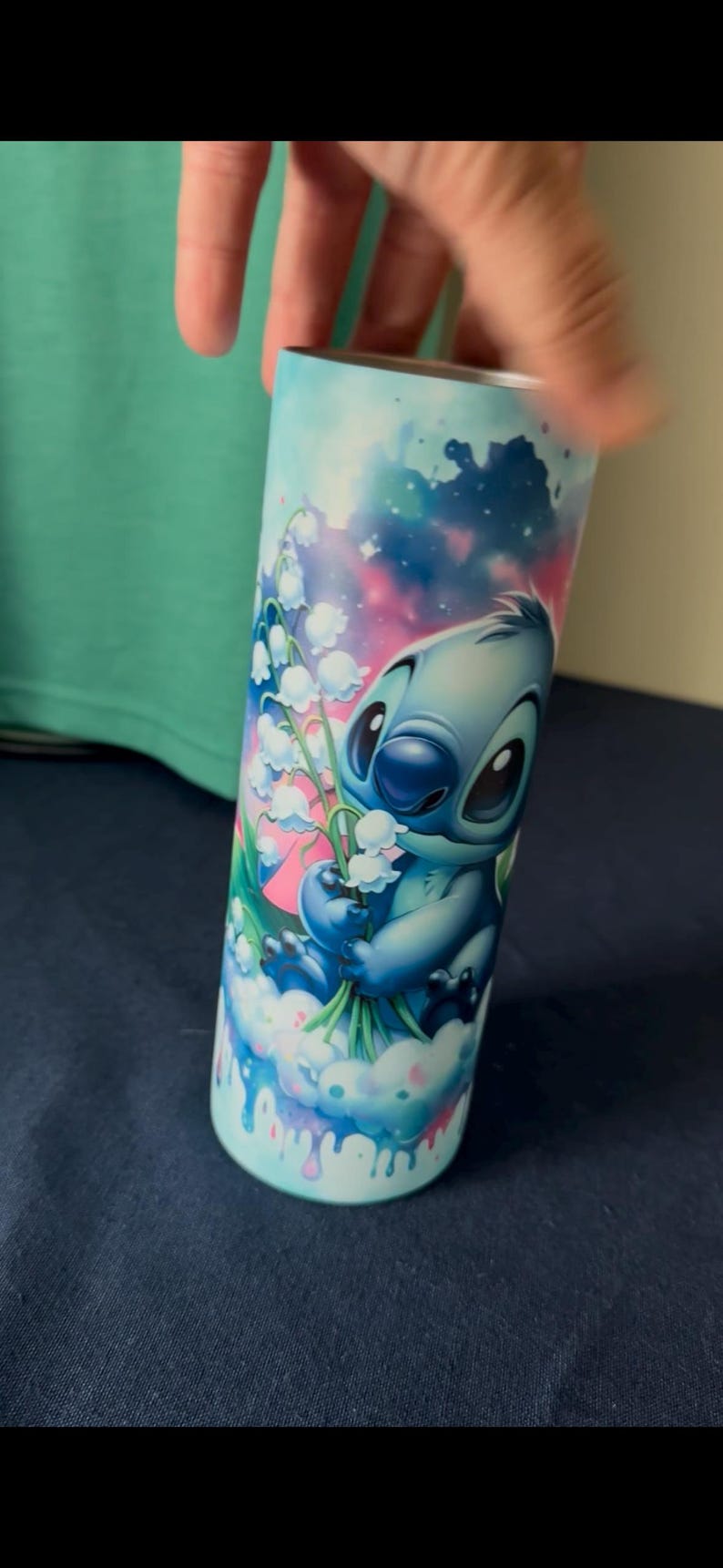May include: A blue tumbler featuring the character Stitch from Lilo & Stitch, holding white flowers. The design includes a sky-blue background with pink and blue accents. The tumbler is cylindrical and appears to be made of metal.