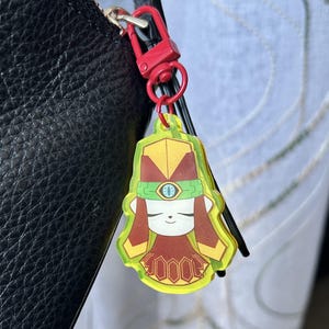 May include: A neon green and red keychain featuring a cartoon character with a yellow headdress, red hair, and a red and brown outfit. The keychain is attached to a red clasp and a black leather bag.