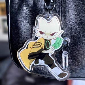 May include: A clear acrylic keychain featuring a cartoon character with yellow and black clothing holding a stack of green bills. The character has white fur and red eyes.