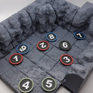 Könnte beinhalten: Ein graues, 3D-gedrucktes Dungeon-Geländeset mit steinartiger Textur. Das Set enthält einen quadratischen Raum mit nummerierten Spielsteinen in Rot, Blau, Schwarz und Grün. Ideal für Tabletop-Gaming.
