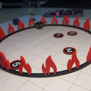 Könnte beinhalten: Ein rundes Spielbrett in Schwarz und Rot mit roten flammenförmigen Teilen am Rand. In der Mitte des Bretts befinden sich vier schwarze und weiße runde Spielsteine mit Zahlen.