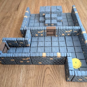 Könnte beinhalten: Ein modulares Dungeon-Gelände-Set mit grauen Steinmauern mit beigen Ziegelakzenten, einer erhöhten Plattform, zwei braunen Holztüren mit schwarzen Scharnieren und einem kleinen gelben runden Teppich. Der Boden besteht aus quadratischen Fliesen.