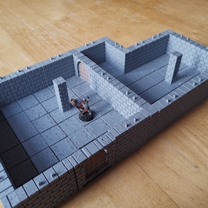 Könnte beinhalten: Ein graues, mit Ziegeln ausgekleidetes Dungeon-Set für Tabletop-Rollenspiele. Das Set enthält zwei Räume mit einer Tür dazwischen und eine Säule in einem der Räume. Das Set ist aus Kunststoff und für die Verwendung mit Miniaturen konzipiert.