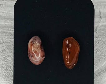 May include: Two polished gemstone earrings on a black display card. One earring is pink and white, the other is reddish-brown. The stones are smooth and rounded. The card has a scalloped top and a hole for display. The earrings are made of natural stone.