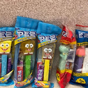 May include: Several PEZ candy dispensers featuring characters from the animated series, SpongeBob SquarePants. The dispensers are in their original packaging, with blue and yellow packaging. The characters include SpongeBob, Squidward, and Patrick.