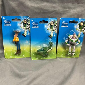 K&ouml;nnte beinhalten: Drei Plastikspielzeugfiguren aus der Disney Toy Story-Reihe. Die Figuren sind Woody, Buzz Lightyear und der Au&szlig;erirdische aus dem Film. Die Figuren sind auf einzelnen Karten mit dem Disney-Logo und den Worten "Disney Figurines" verpackt.