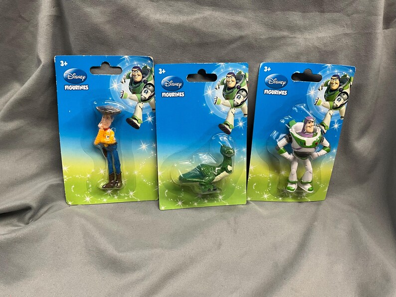 K&ouml;nnte beinhalten: Drei Plastikspielzeugfiguren aus der Disney Toy Story-Reihe. Die Figuren sind Woody, Buzz Lightyear und der Au&szlig;erirdische aus dem Film. Die Figuren sind auf einzelnen Karten mit dem Disney-Logo und den Worten "Disney Figurines" verpackt.