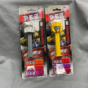 May include: Two Transformers character Pez dispensers, one gray and one yellow, with a pack of Pez candies. The dispensers are on a white background with a blue and white Transformers logo.