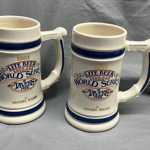 May include: Two white ceramic beer steins with blue accents and handles. Each stein features the text "1983 Lite Beer World Series of Tavern Pool at Caesars Palace" in red, blue, and gold lettering.