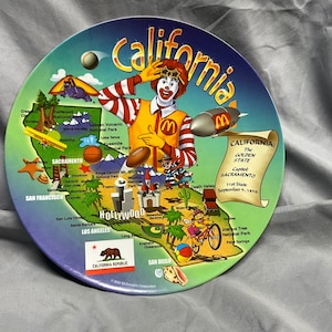 May include: A colorful ceramic plate featuring a map of California with various landmarks and attractions. The plate also includes a cartoon character dressed as Ronald McDonald, waving and holding a golden M. The text "California: The Golden State" is written on a scroll at the top of the plate.