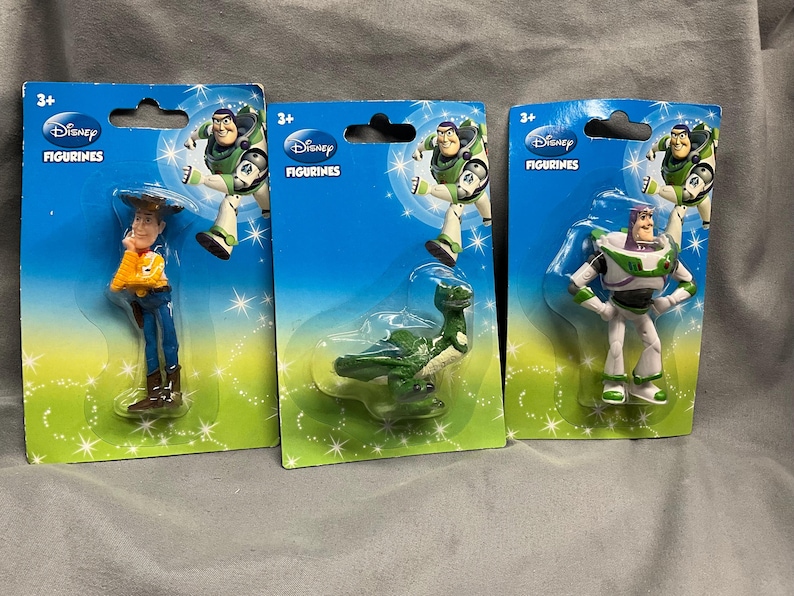 K&ouml;nnte beinhalten: Drei Plastik-Disney-Figuren auf blauem Hintergrund. Die Figuren sind Buzz Lightyear, Woody und Rex. Die Verpackung tr&auml;gt den Schriftzug "Disney Figuren".