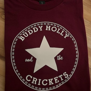 Puede incluir: Una camiseta color granate con un gráfico blanco de una estrella dentro de un círculo. El círculo tiene el texto "Buddy Holly and the Crickets" alrededor de la estrella.