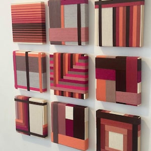 Op de afbeelding: Negen vierkante wandkunstwerken met abstracte ontwerpen. Elk stuk heeft een uniek patroon van gelaagd, kleurrijk garen in tinten rood, oranje, roze en bruin. Het kunstwerk is op een witte muur gemonteerd en creëert een moderne, geometrische weergave.