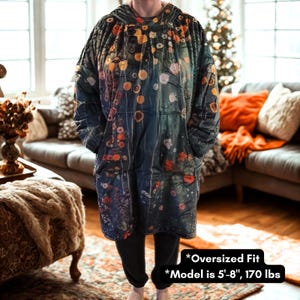 K&ouml;nnte beinhalten: Eine &uuml;bergro&szlig;e Kapuzen-Decke mit einem Blumenmuster in Blau-, Gr&uuml;n-, Orange- und Gelbt&ouml;nen. Die Decke hat eine gro&szlig;e Vordertasche und eine Kapuze. Der Text auf dem Bild lautet "Oversized Fit" und "Model ist 173 cm, 77 kg."