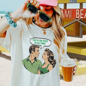 May include: Cream-colored t-shirt featuring a retro comic-style illustration of a man and woman. The woman is speaking the phrase, "You're the Pivot to my Table" in a speech bubble. The shirt is worn by a person holding sunglasses and a beverage.