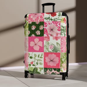 Pode incluir: Uma mala preta com rodas com um design de patchwork floral. O padrão apresenta quadrados rosa, verde e branco com vários motivos de flores e folhas. A mala tem uma alça retrátil e quatro rodas pretas.