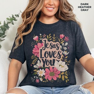 May include: A dark heather grey t-shirt with a floral design and the text "Jesus Loves You" in pink.