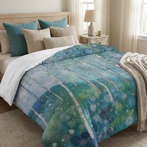 May include: A bed with a comforter featuring a watercolor design of birch trees in shades of blue, green, and white. Teal and beige pillows are arranged on the bed, along with a beige throw blanket. The bed is in a well-lit room.